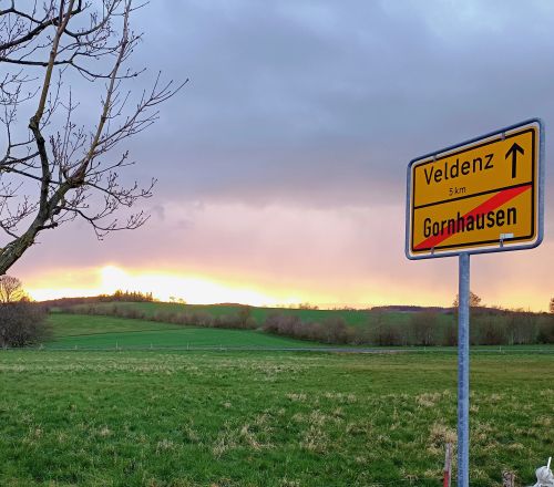 Ortsschild Veldenz in der Landschaft mit Wiese Baum und Sonnenuntergang im Hintergrund