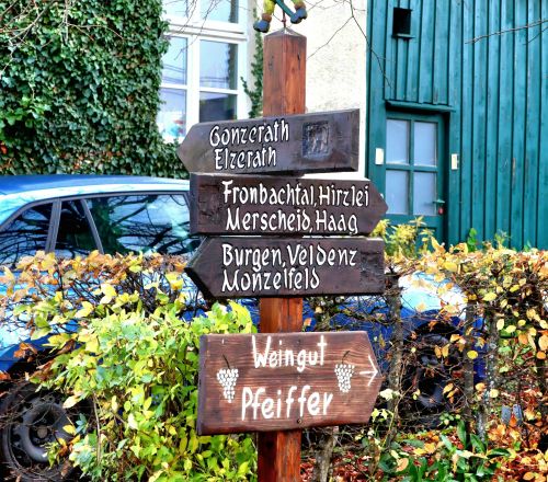 Holzwegweiser mit mehreren Ortsschildern und Figur eines Wanderers vor Haus mit blauen Türen und grünem Auto