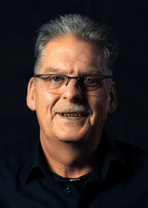 Porträt eines lächelnden älteren Mannes mit grauen Haaren Brille und schwarzem Hemd vor dunklem Hintergrund